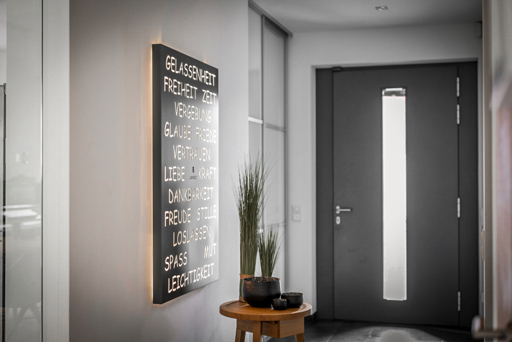 Ein dunkles Kraft-Licht-Werk an der linken Wand im Eingangsbereich. Gerade aus ist eine dunkelgraue Eingangstür.